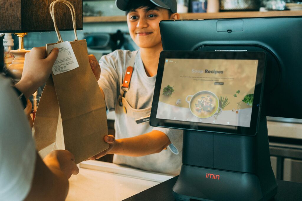 pexels-photo-12935094-12935094 A smiling cashier hands a paper bag to a customer at a modern point of sale, showcasing customer service.